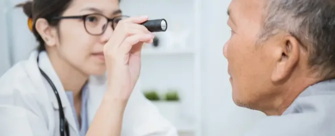 A medical professional holds a flashlight towards the eye of an elderly male patient during an eye exam, checking for any abnormalities or vision issues