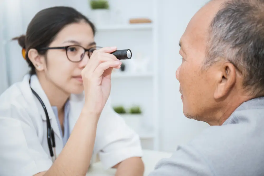 A medical professional holds a flashlight towards the eye of an elderly male patient during an eye exam, checking for any abnormalities or vision issues