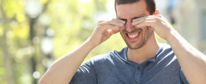 A man squinting and rubbing his eyes, standing outdoors in bright sunlight, suggesting discomfort or sensitivity to light