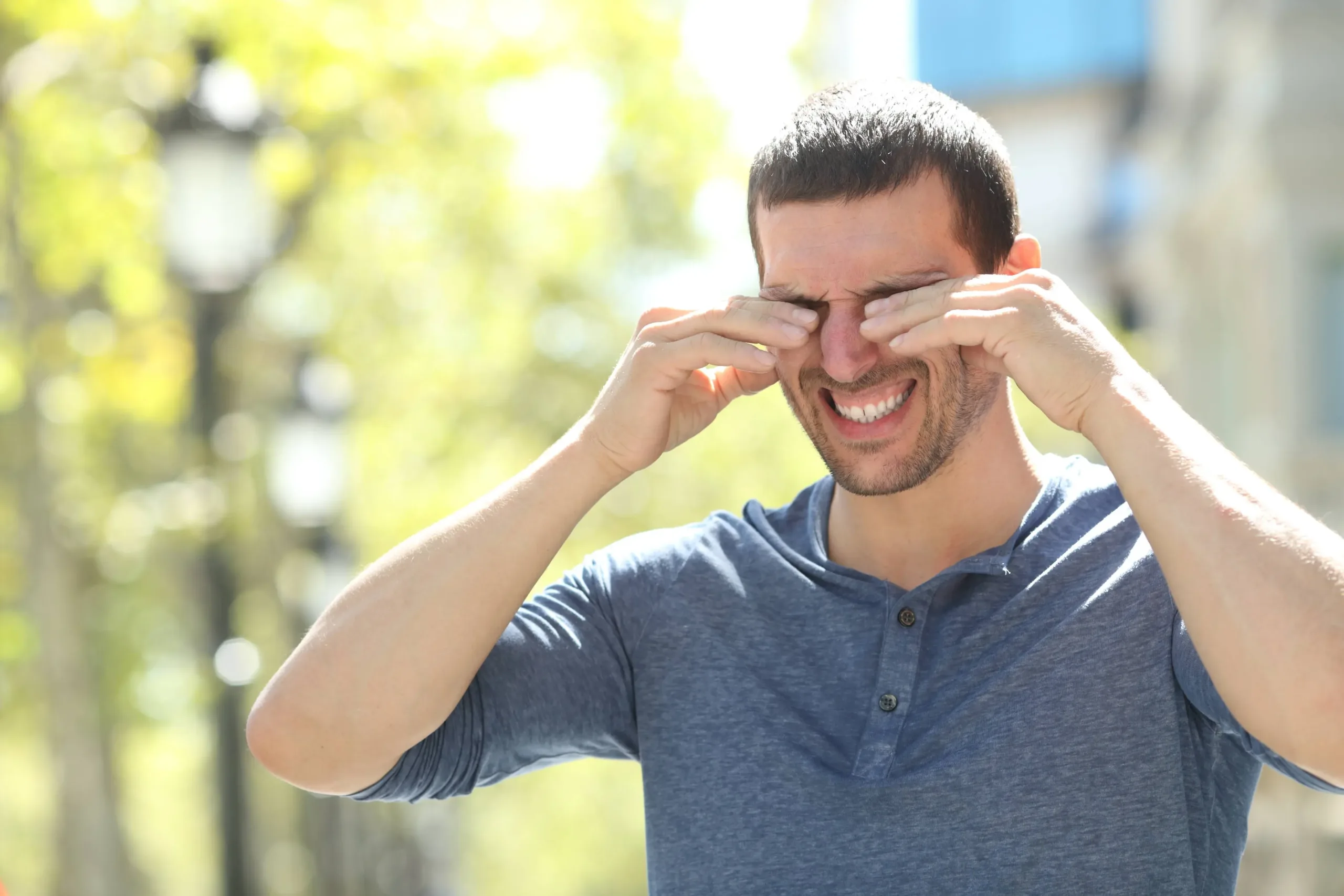 A man squinting and rubbing his eyes, standing outdoors in bright sunlight, suggesting discomfort or sensitivity to light