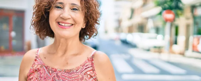 A woman smiling brightly at the camera while outdoors, exuding a cheerful and positive vibe against an urban backdrop