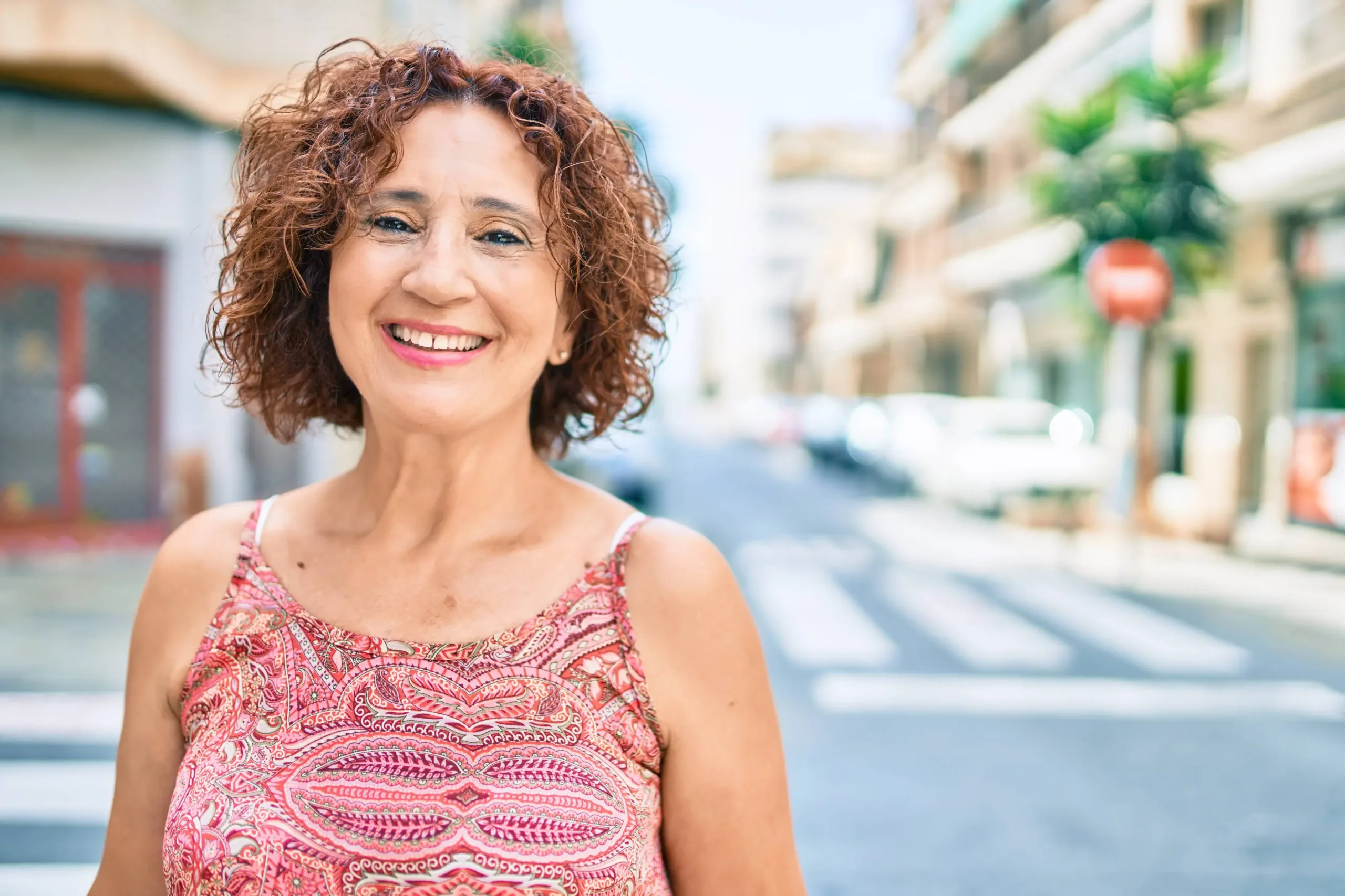 A woman smiling brightly at the camera while outdoors, exuding a cheerful and positive vibe against an urban backdrop