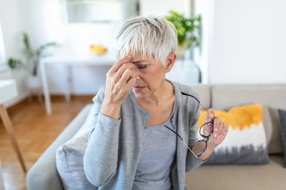 A woman appears irritated as she holds her nose with a frustrated expression, having removed her eyeglasses