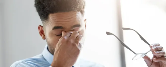 A man appears irritated as he holds his nose with a frustrated expression, having removed his eyeglasses