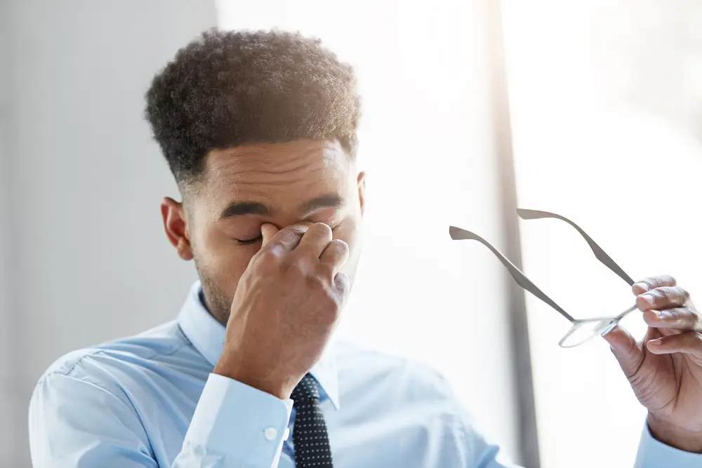 A man appears irritated as he holds his nose with a frustrated expression, having removed his eyeglasses